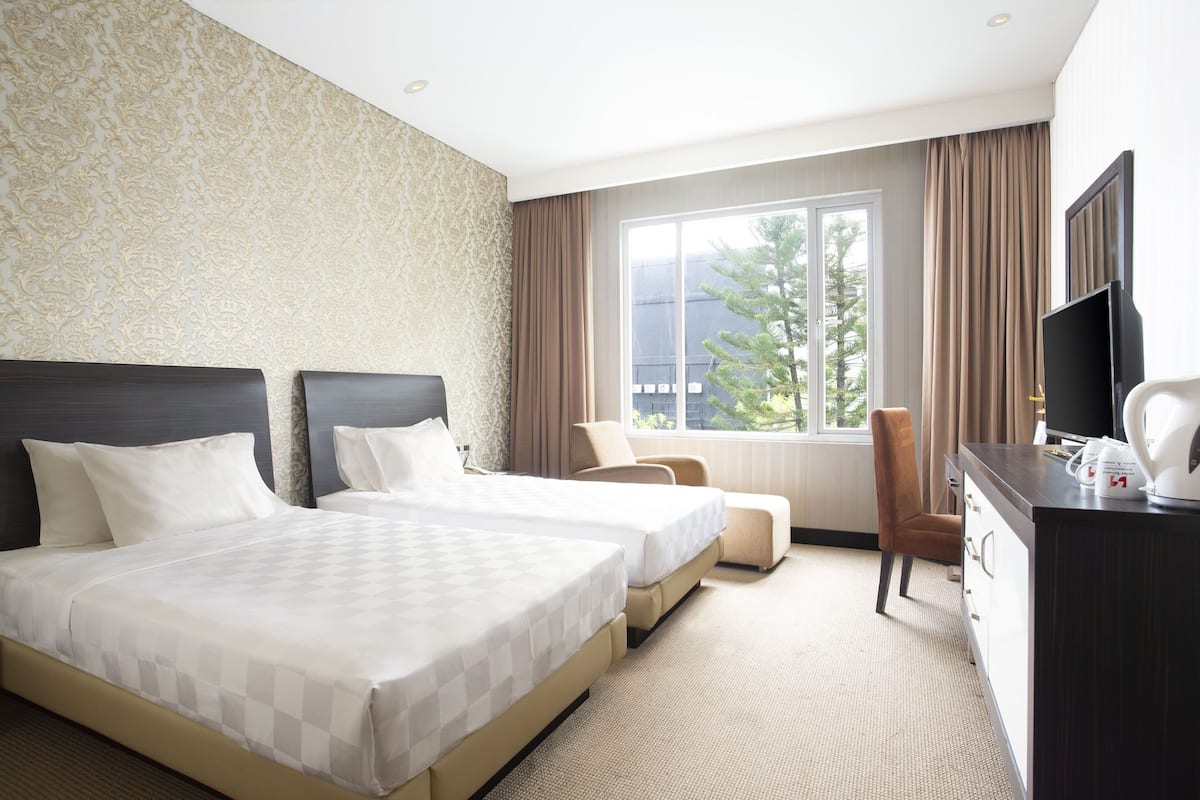 A hotel room with two beds, a desk, a chair, and a large window with a view of trees.
