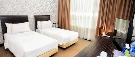 A hotel room with two beds, a wooden desk, a television, and a window with curtains.