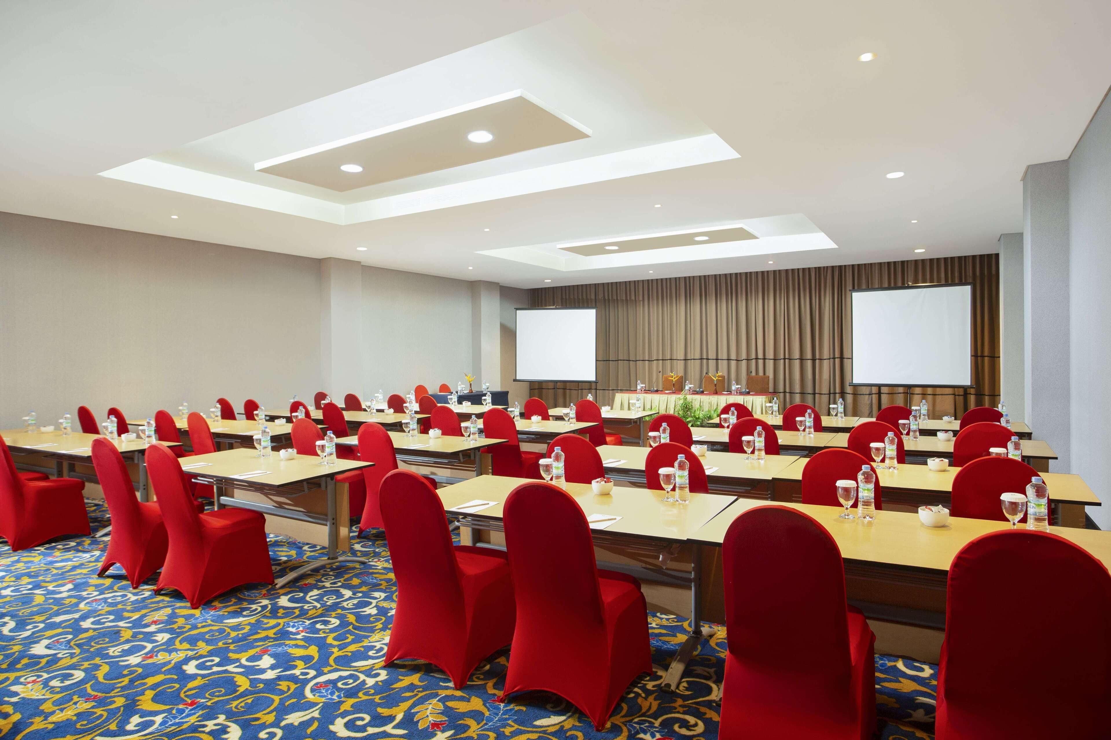 A conference room with rows of tables and red chairs, equipped with water bottles and glasses, and a large screen at the front.