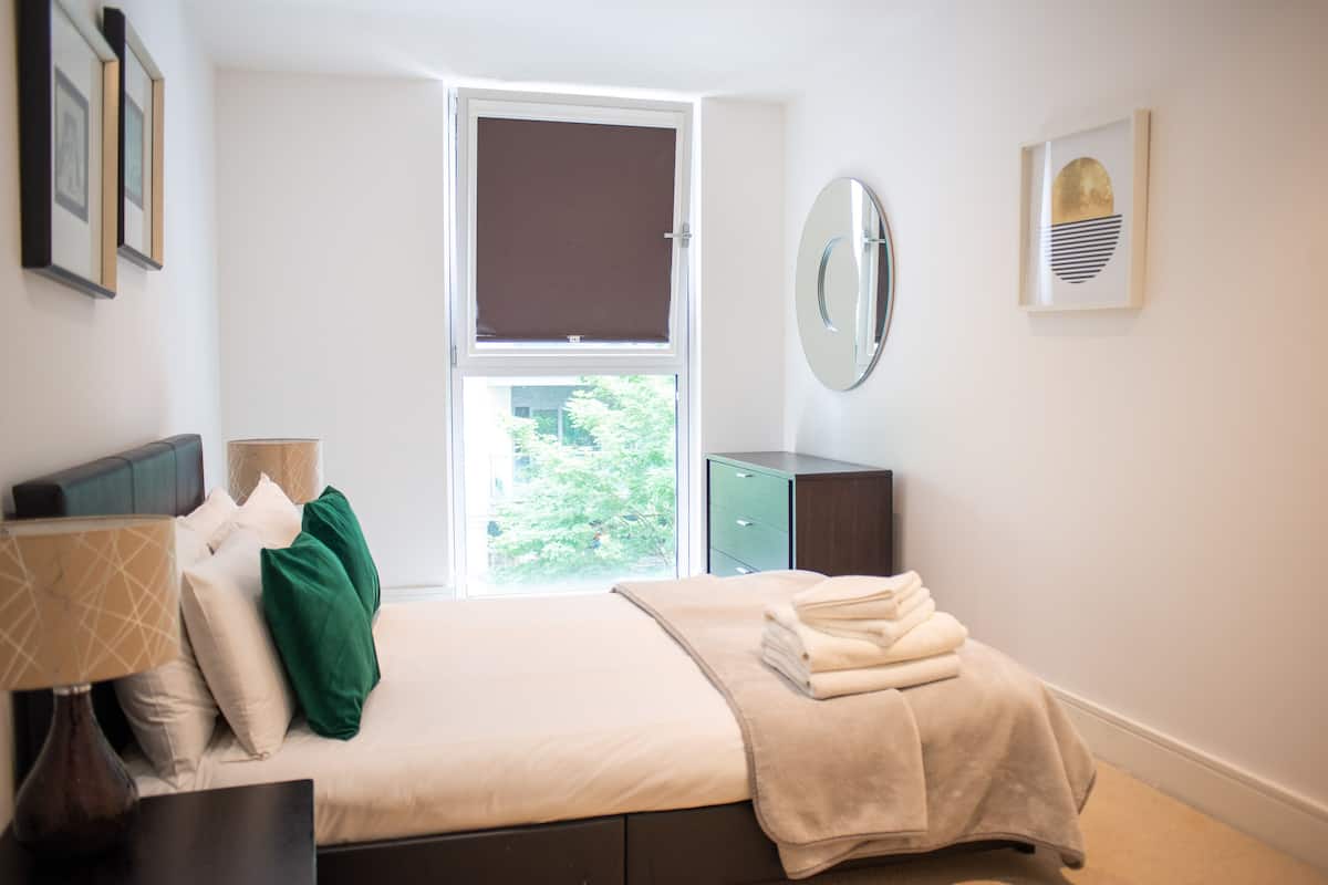 A modern bedroom with a bed, bedside table, lamp, and a window with blinds.