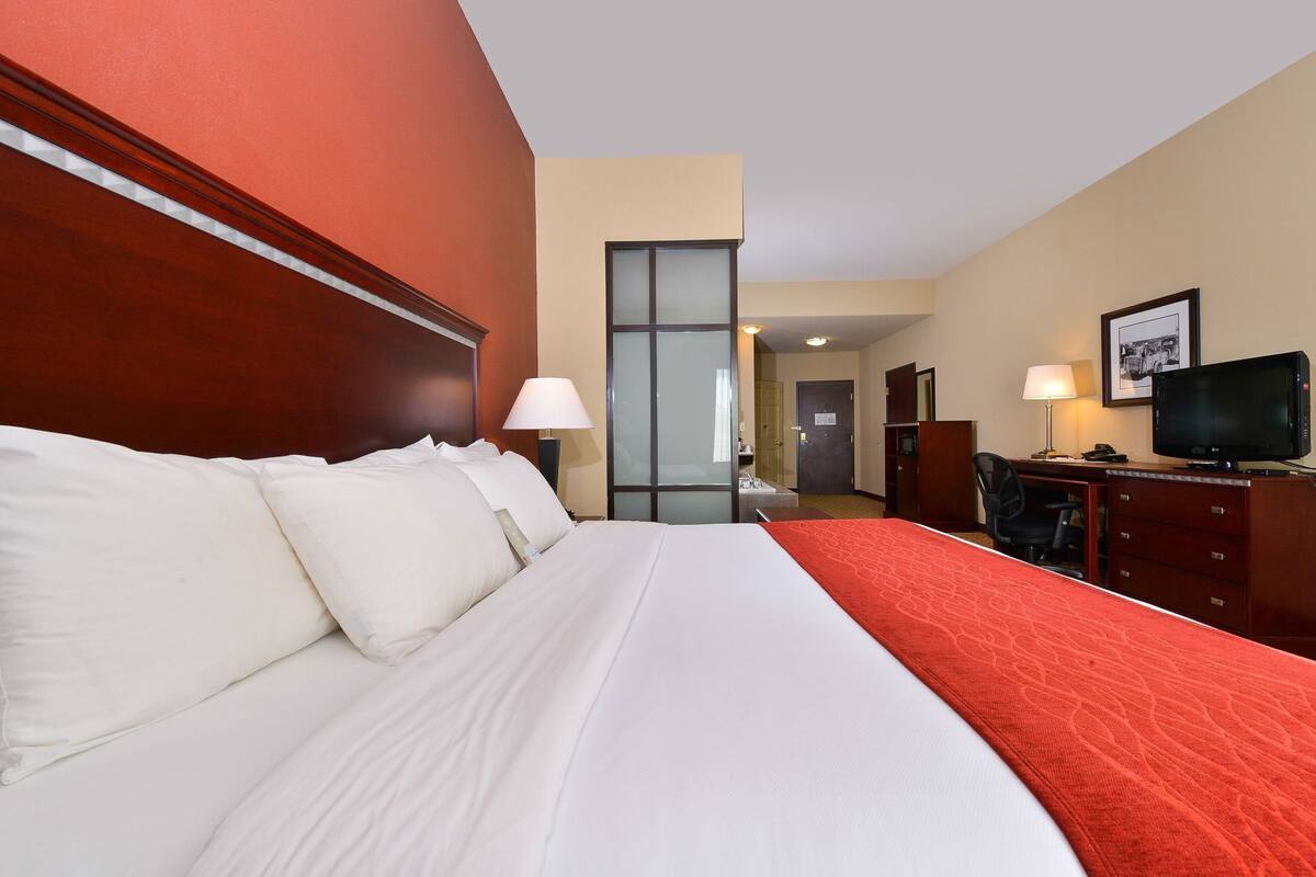A hotel room with a large bed, a desk with a television, and a bathroom visible in the background.