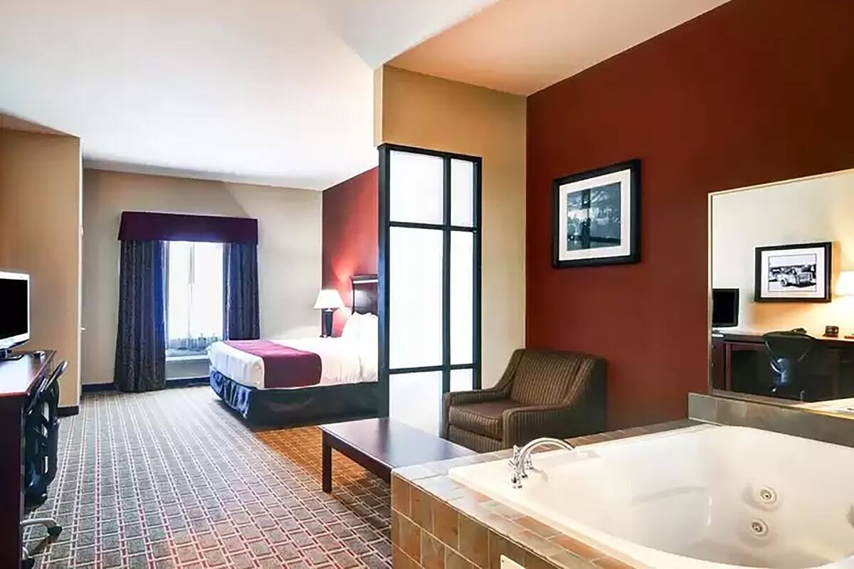 Ett hotellrum med en säng, en stol, ett skrivbord, en tv, ett stort fönster och ett badkar.