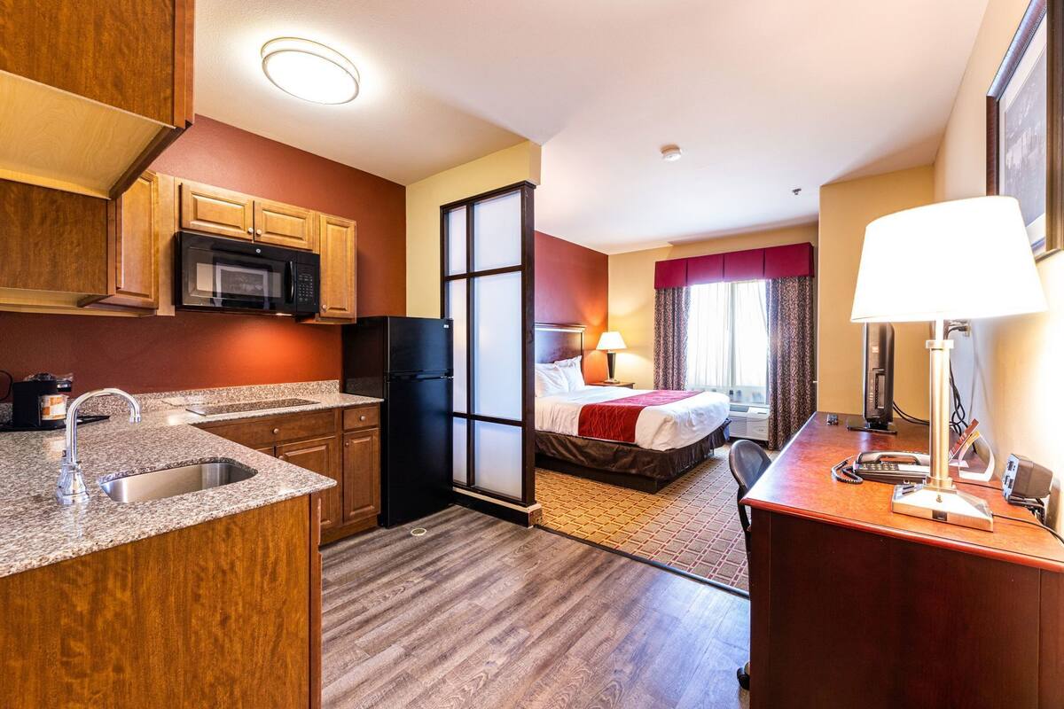 A hotel room with a kitchenette, a bed, a desk with a lamp, and a phone.