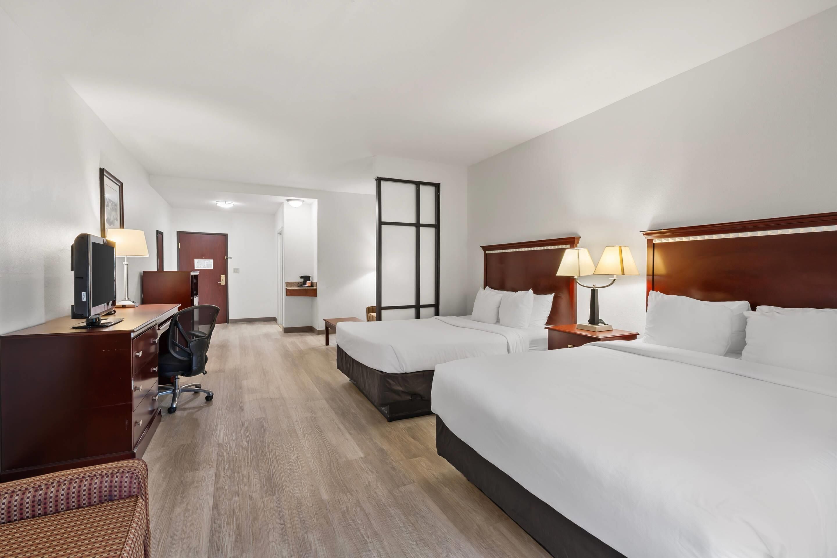 A hotel room with two beds, a desk with a computer, a chair, and a small table with a lamp.