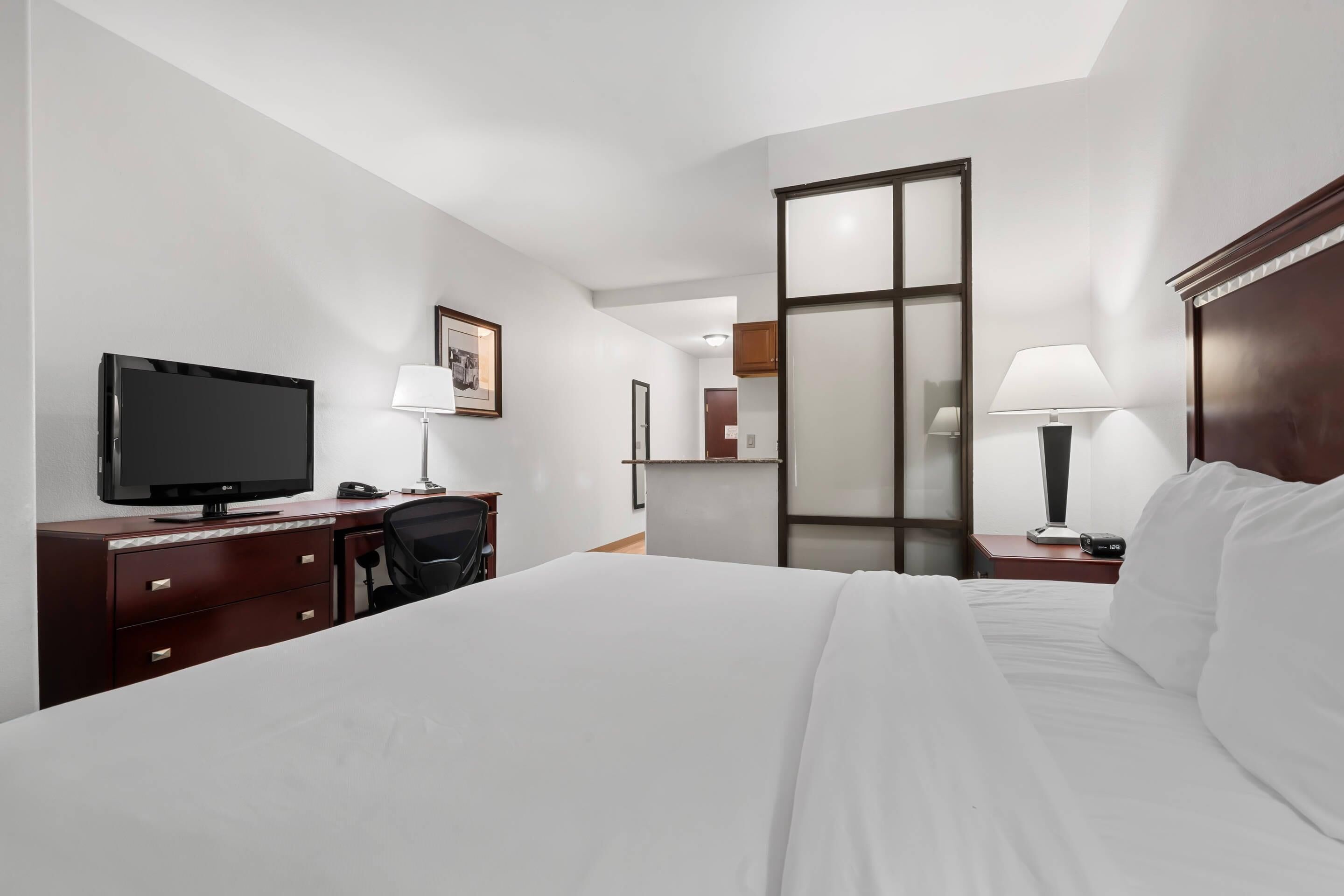 Ein Hotelzimmer mit einem Bett, einem Schreibtisch mit Fernseher, einem Sessel, einem Nachttisch mit Lampe und einem Spiegel.