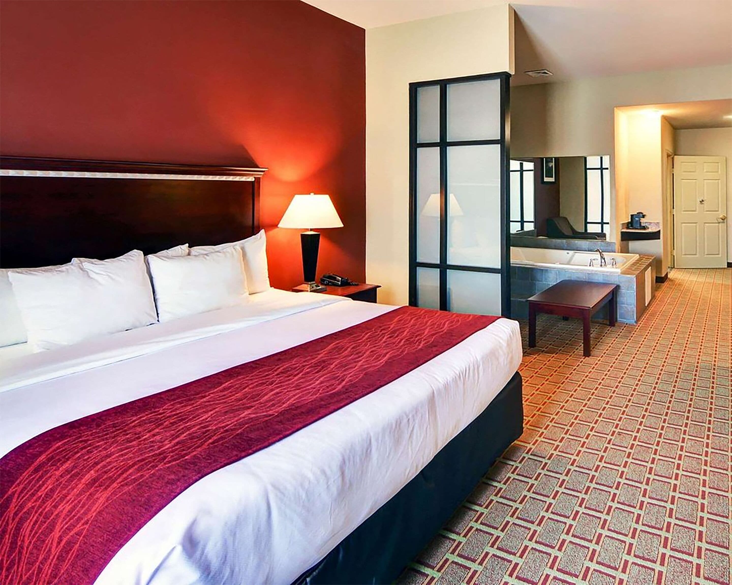 Habitación de hotel con una cama grande, una mesita de noche con lámpara, un baño con lavamanos y espejo, y una alfombra estampada.
