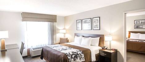 A hotel room with a large bed, bedside tables, a desk, and a chair. There are framed pictures on the wall and a window with curtains.