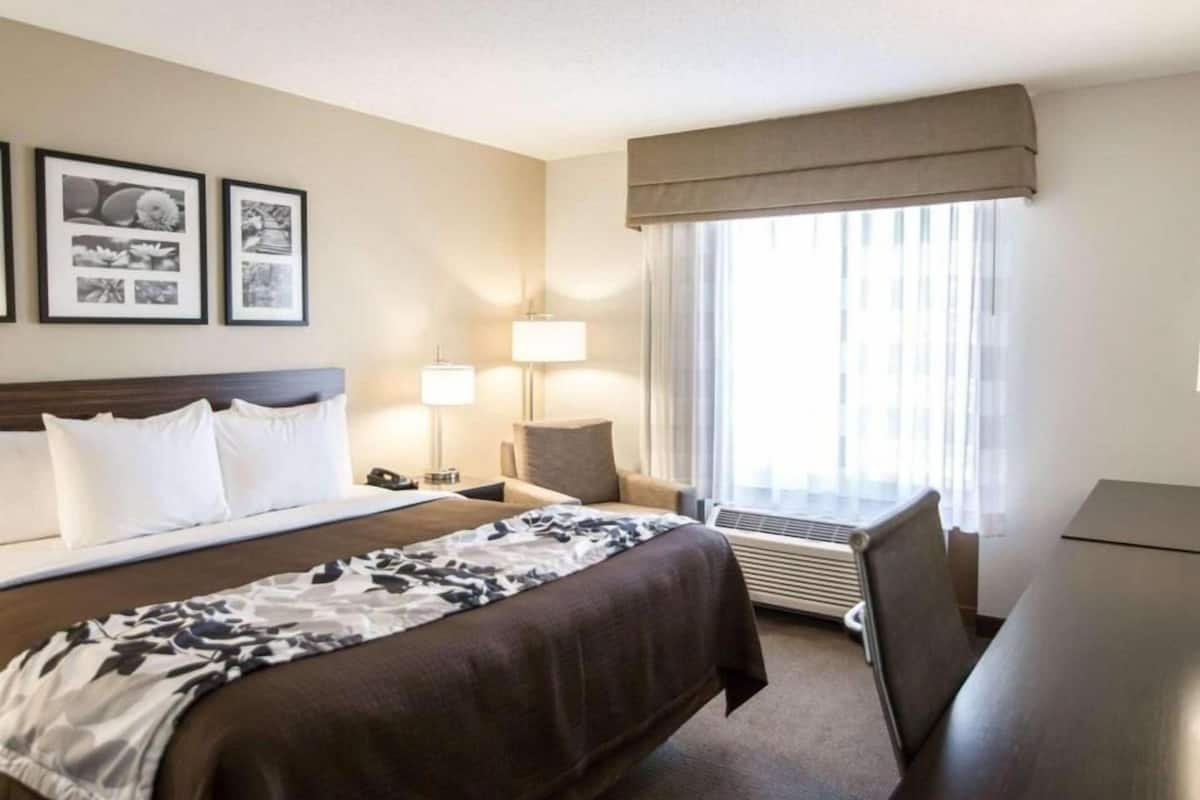 A hotel room with a large bed, bedside tables, a desk, a chair, a window with curtains, and framed pictures on the wall.