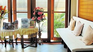 A room with a sofa, a table with magazines, a vase with flowers, and a view of greenery outside. - LaOr Inn Hua Hin (Hua Hin)