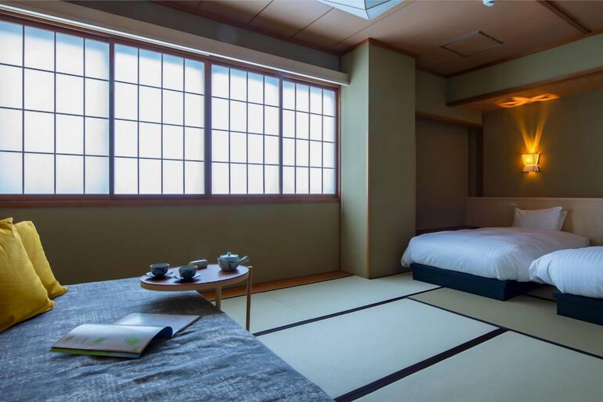 A hotel room with tatami flooring, a bed, a sofa, a small table with tea set, and a wall-mounted light.