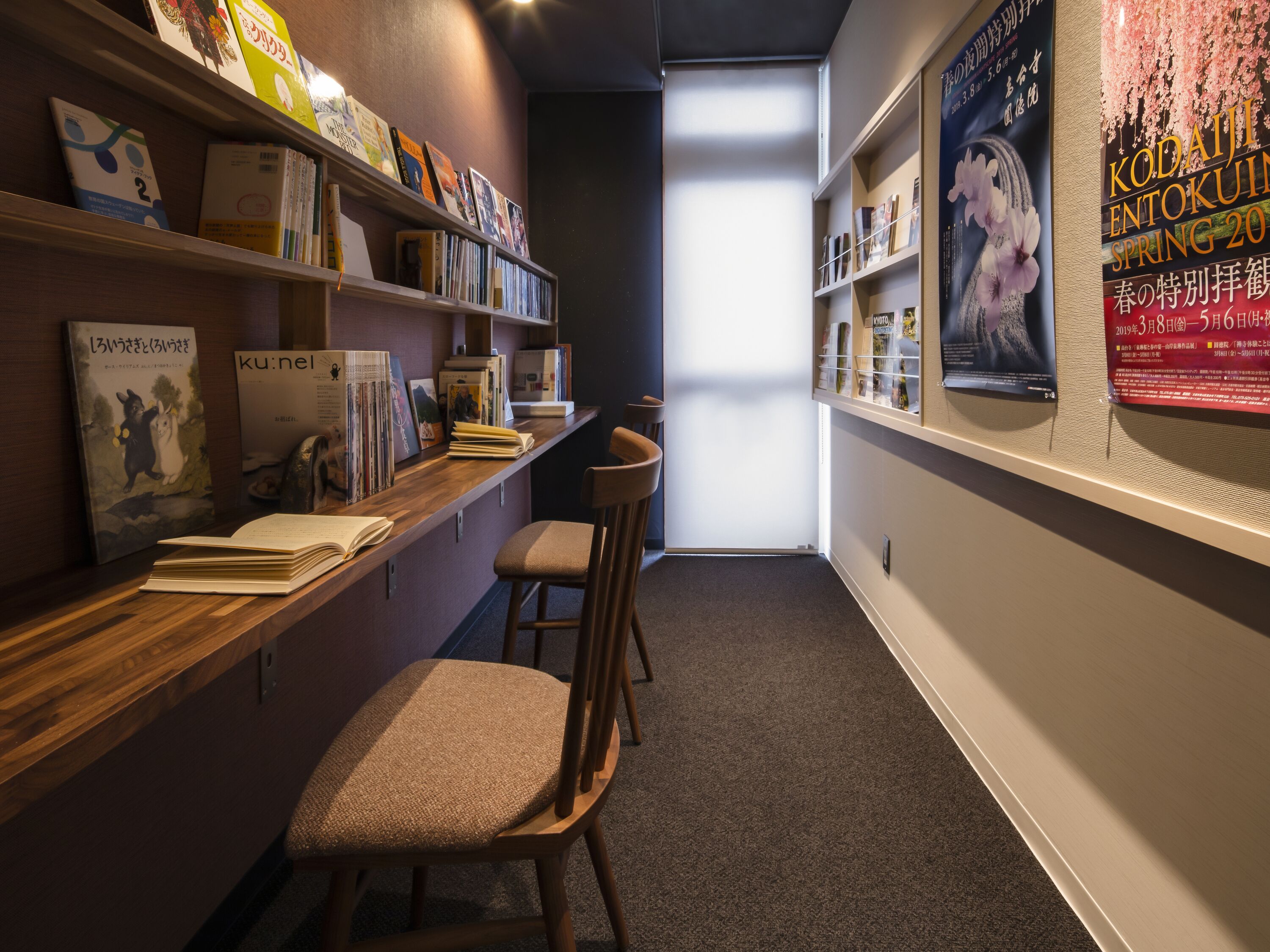 A narrow hallway with bookshelves on both sides, a wooden desk, and two bar stools.