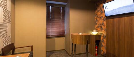 Habitación de hotel con una mesa de madera, una silla, una mesita redonda con lámpara, un televisor montado en la pared y una ventana con persianas.