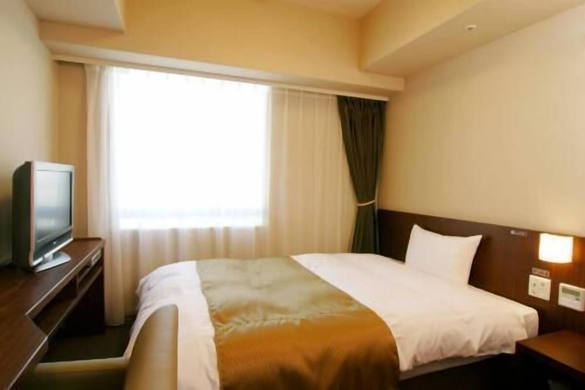 Hotellihuone, jossa on sänky, jalustalla oleva televisio, ikkuna, jossa on verhot, ja seinälle asennettu valaisin.