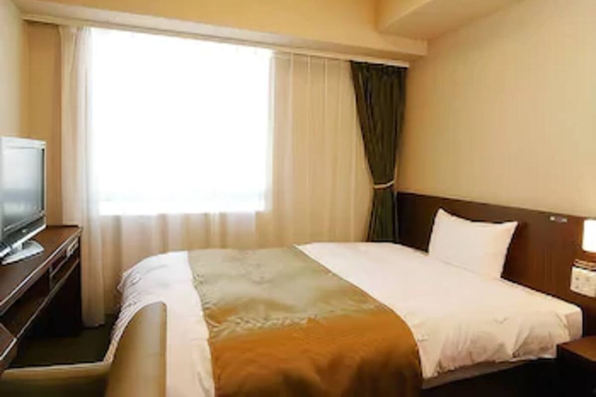 Hotellihuone, jossa on sänky, jalustalla oleva televisio, ikkuna, jossa on verhot, ja yöpöytä, jossa on lamppu.