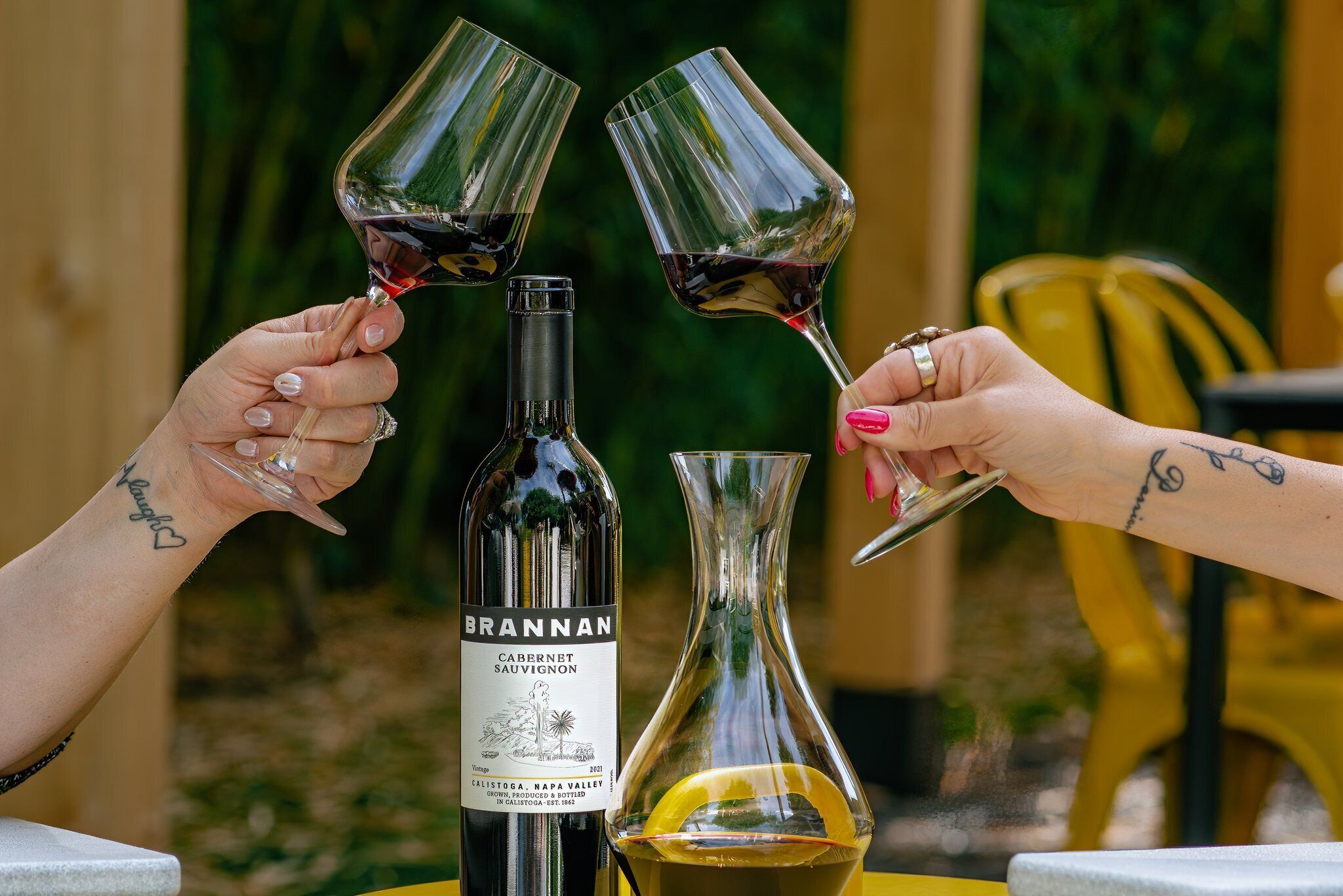 A bottle of Brannan Cabernet Sauvignon and a glass decanter on a table with two people toasting with wine glasses.