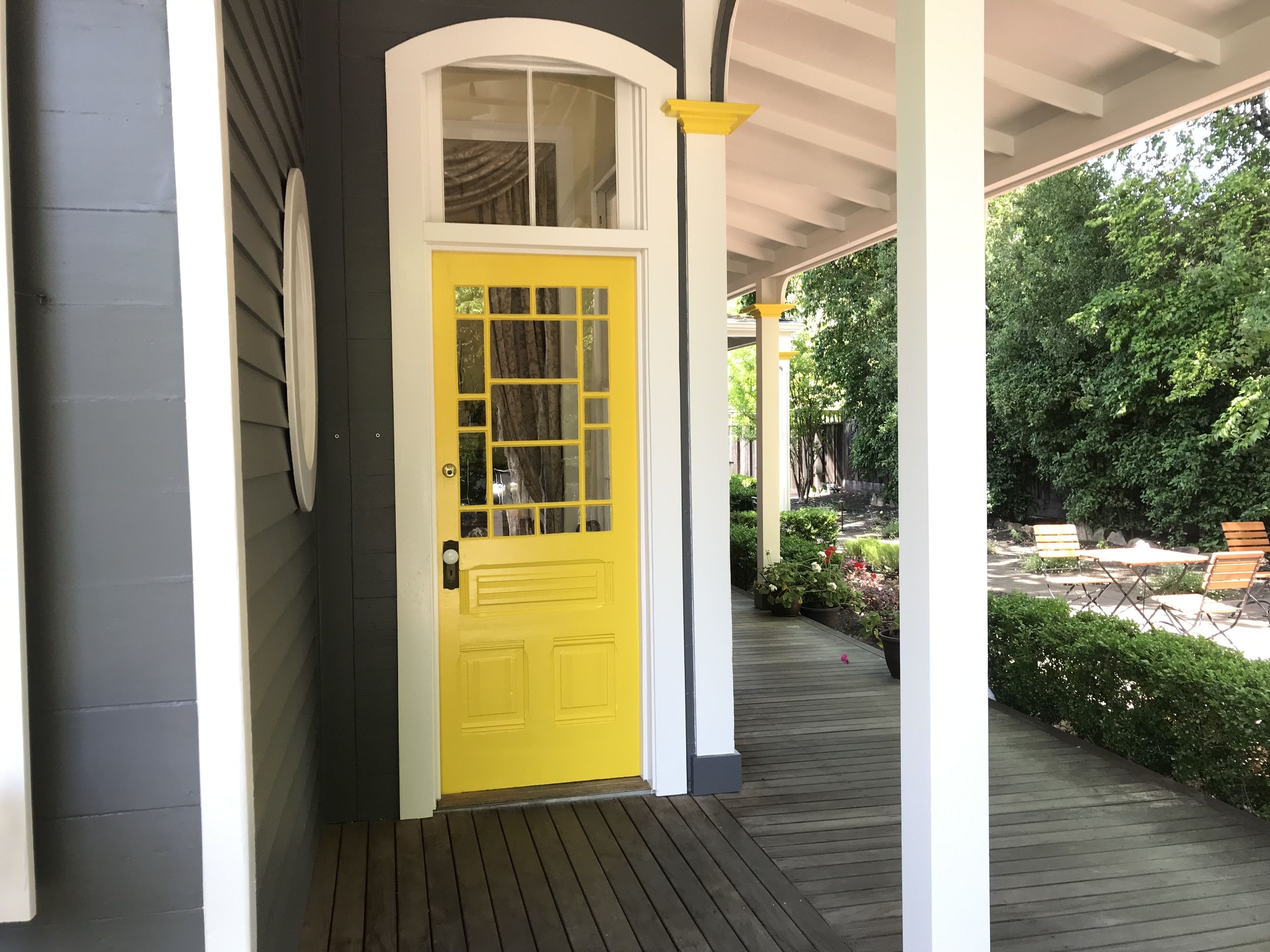 Una puerta amarilla con ventana, un porche cubierto con suelo de madera y un jardín con bancos.