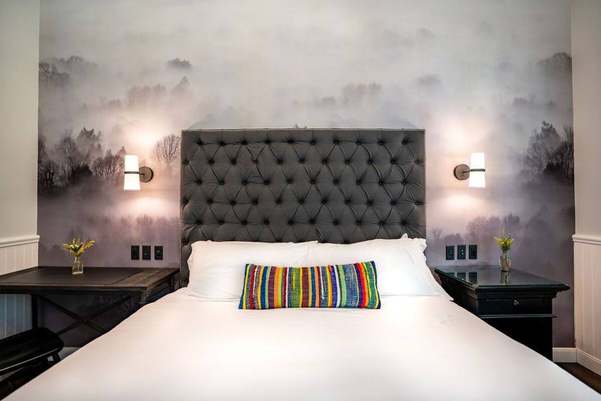 Une chambre à coucher avec une tête de lit capitonnée, du linge de lit blanc et un coussin rayé aux couleurs vives. On y trouve deux tables de chevet avec des lampes et de petits vases ornés de fleurs.