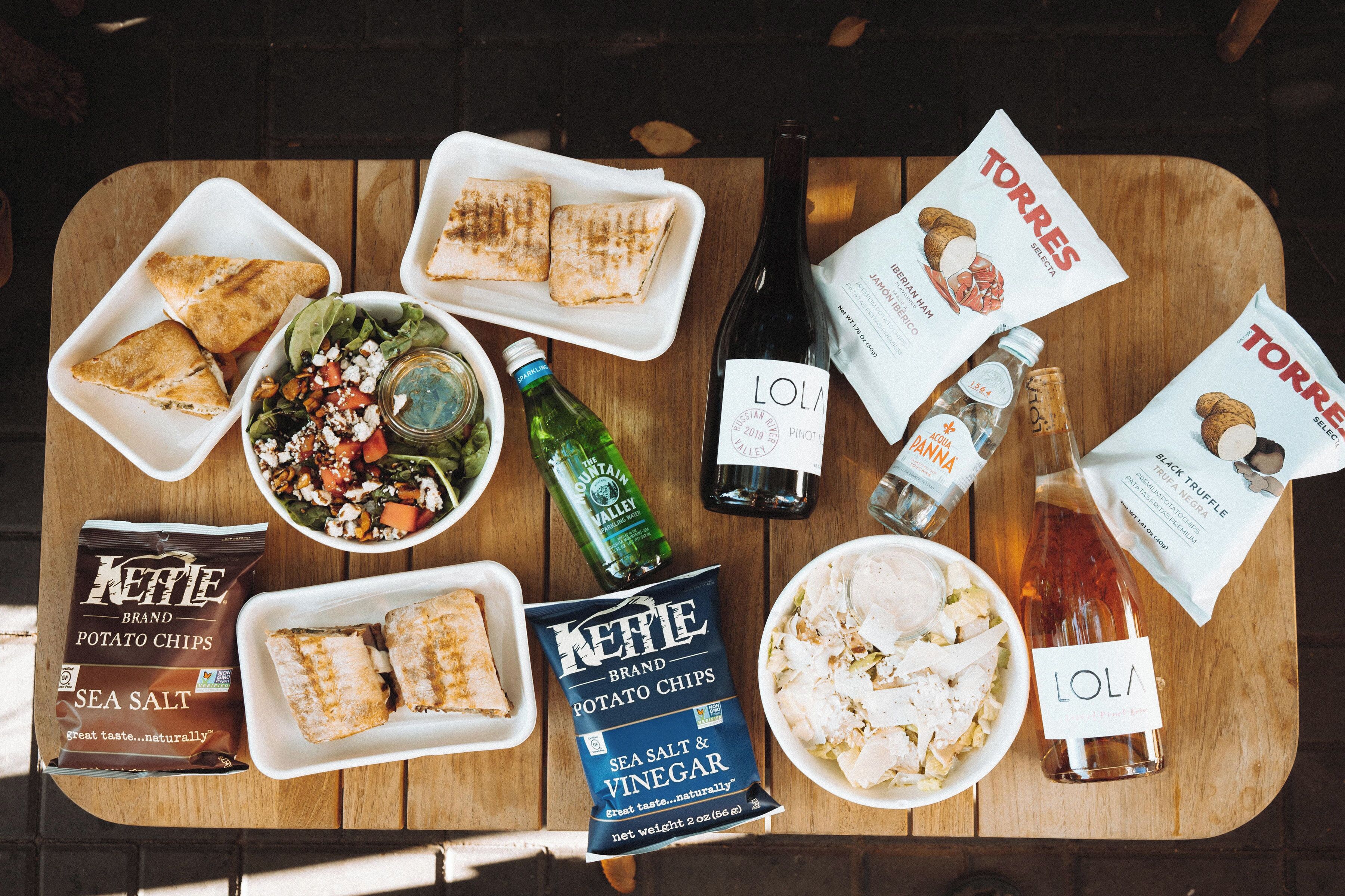 A wooden table with various food items, including sandwiches, salads, and potato chips, along with bottles of wine and a bottle of water.
