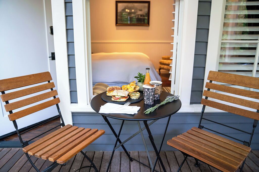 Ein kleiner Sitzbereich im Freien mit zwei Holzstühlen und einem Tisch, einer Frühstücksauslage und Blick auf ein Schlafzimmer durch ein Fenster.