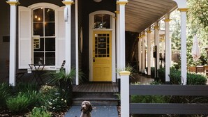 Una casa con puerta amarilla y columnas blancas, un perro sentado adelante y un porche cubierto.