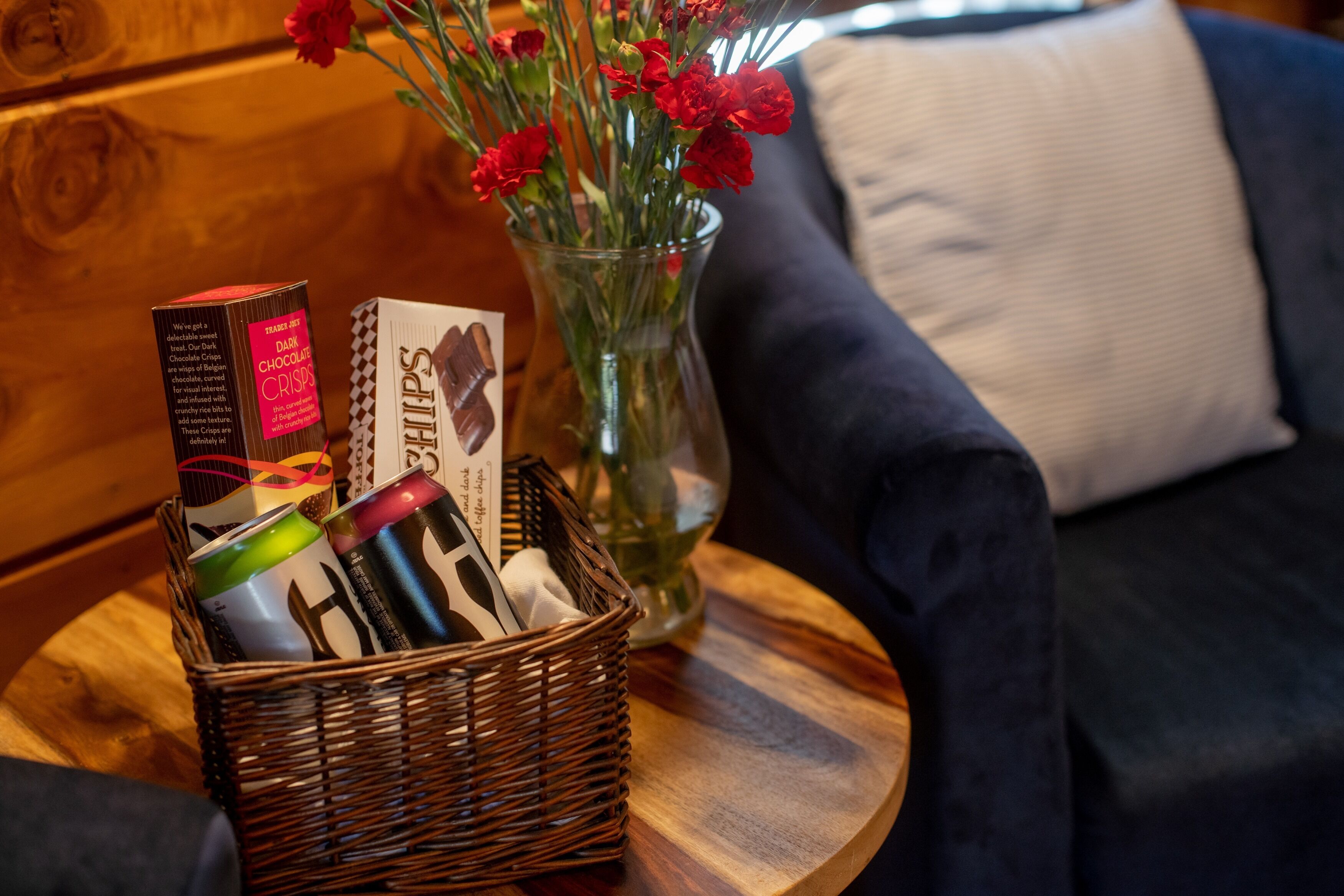 Una cesta de mimbre con bocadillos y una caja de bombones sobre una mesa de madera junto a un florero con flores rojas.