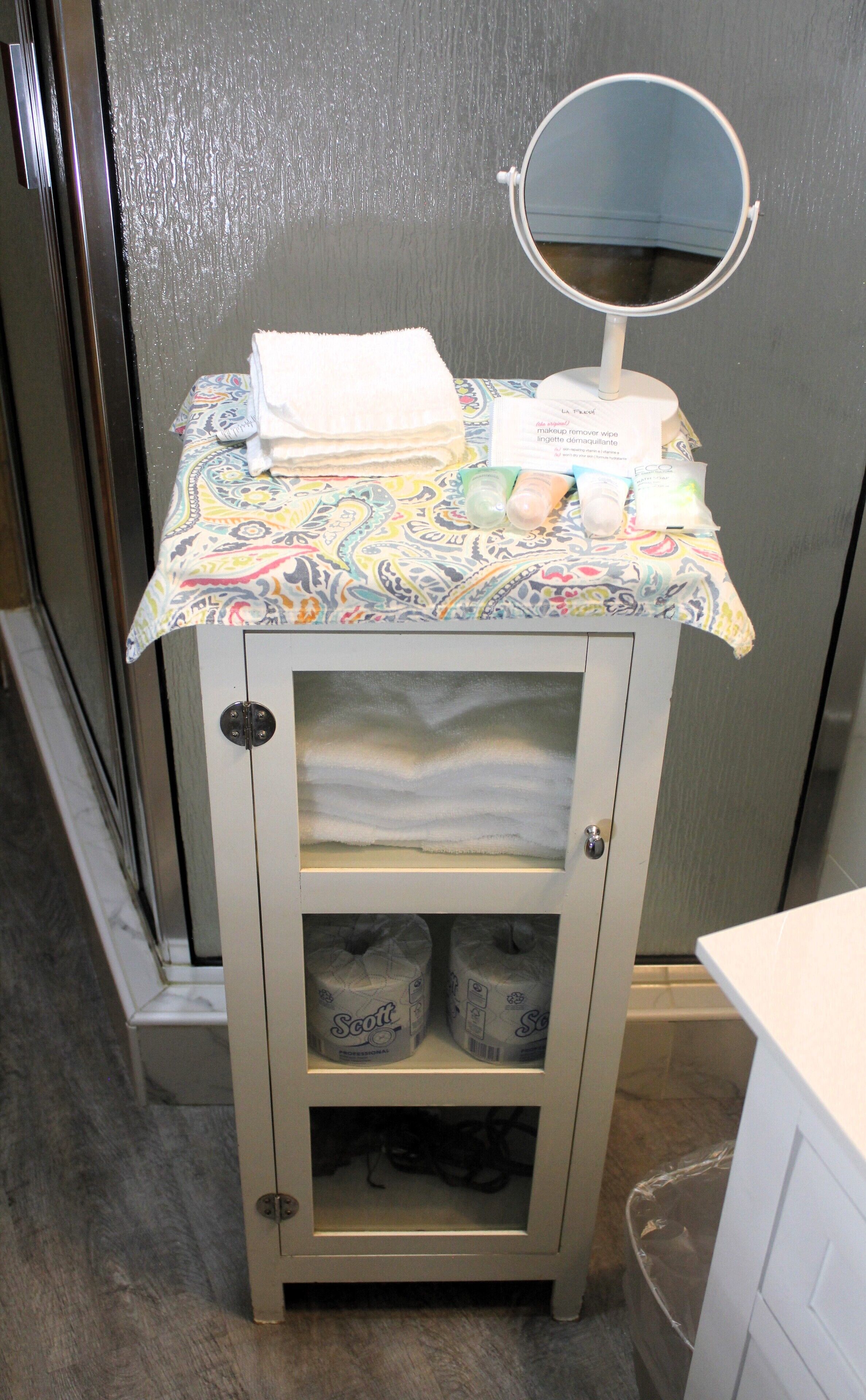 A bathroom vanity with a patterned tablecloth, a folded towel, a makeup remover wipe, and two rolls of Scott toilet paper.