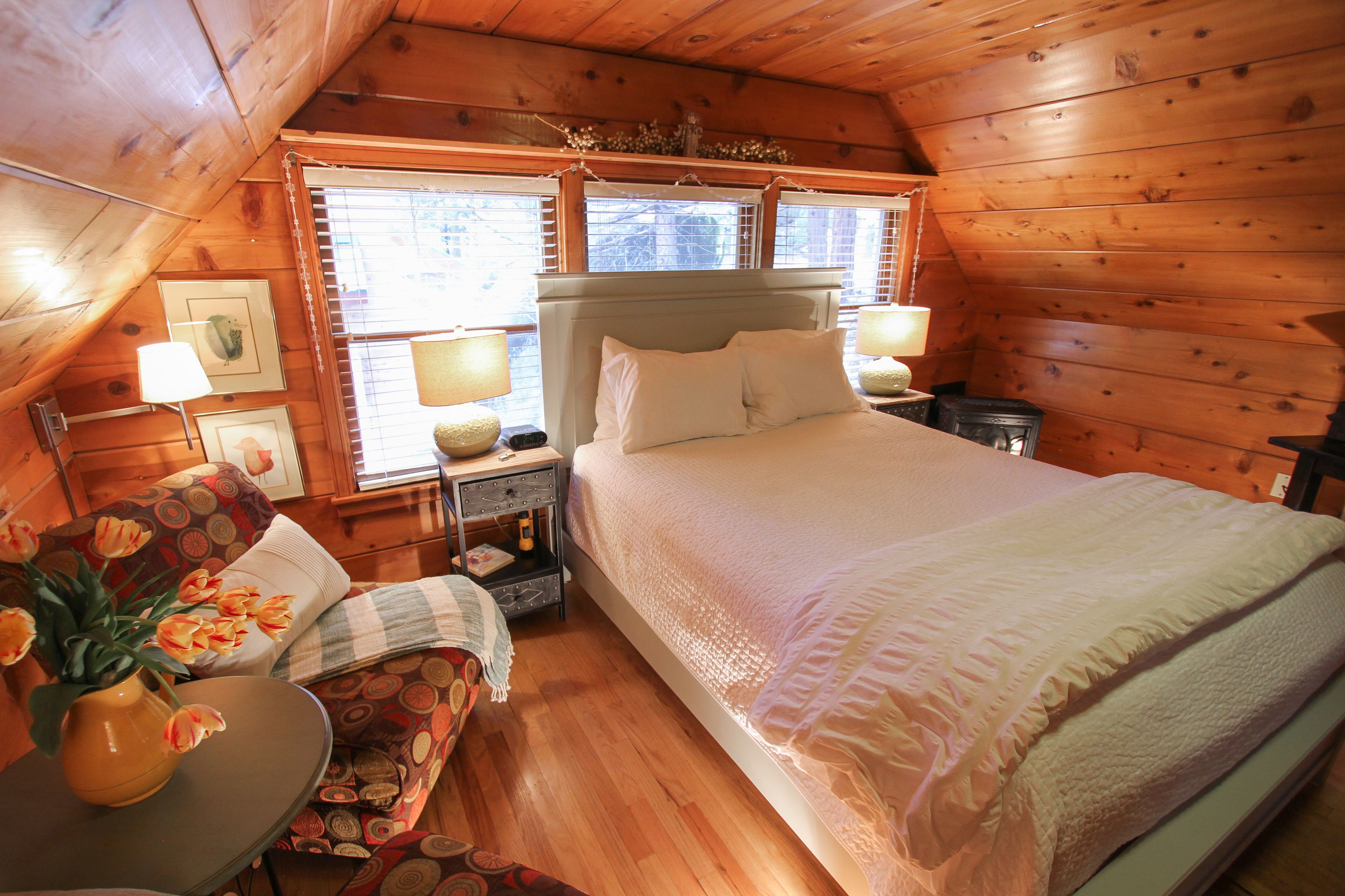 A cozy wooden cabin bedroom with a large bed, a nightstand with a lamp, a small table with a vase of flowers, and a window with blinds.