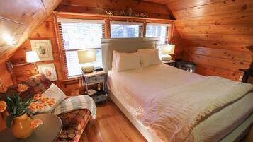 A cozy wooden cabin bedroom with a large bed, a nightstand with a lamp, a small table with a vase of flowers, and a window with blinds.