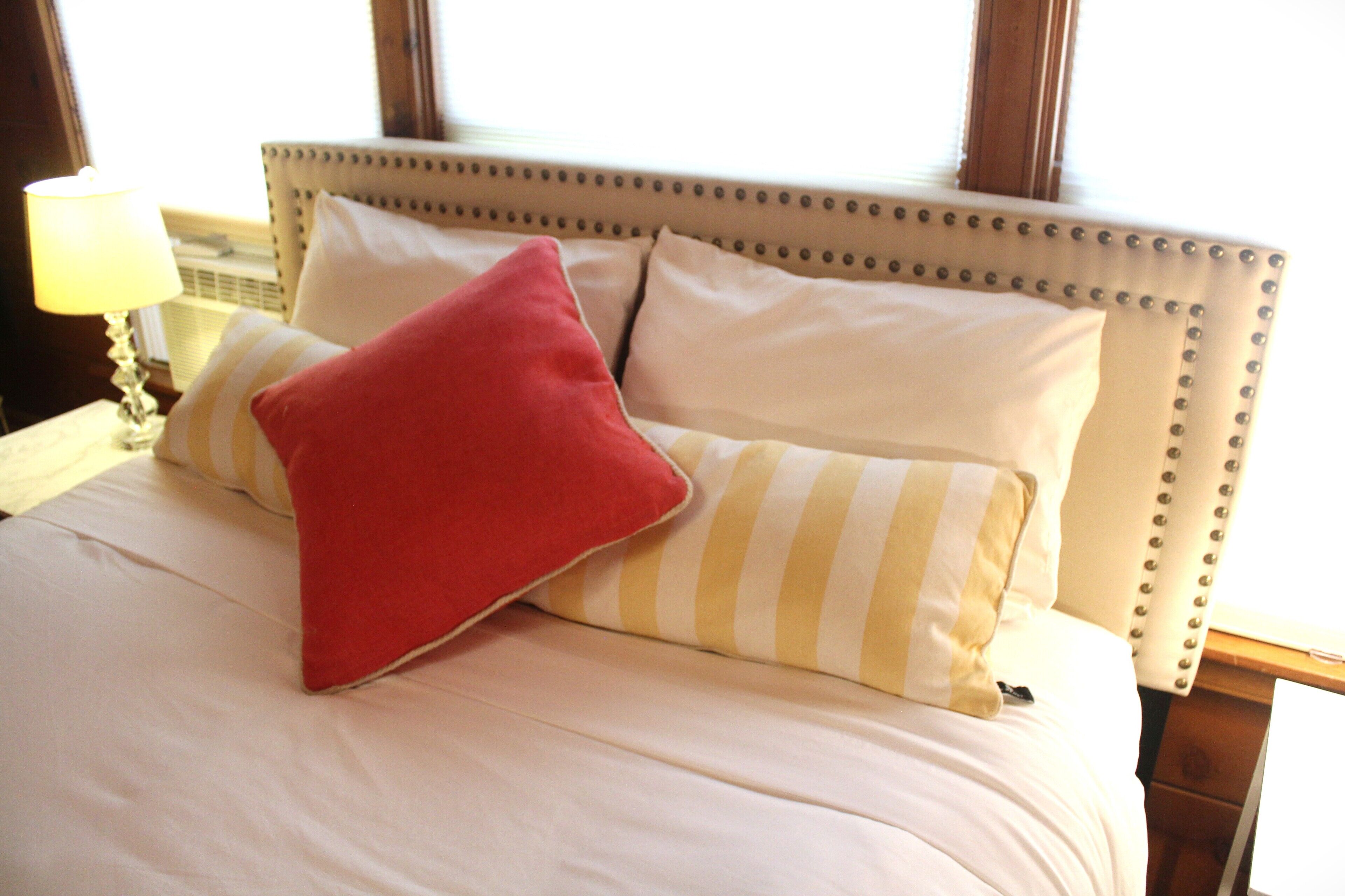A bed with a striped pillow and a red pillow, a lamp, and a window in the background.