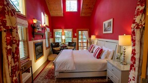 A bedroom with a large bed, a fireplace, a desk, and a window with a view of trees.