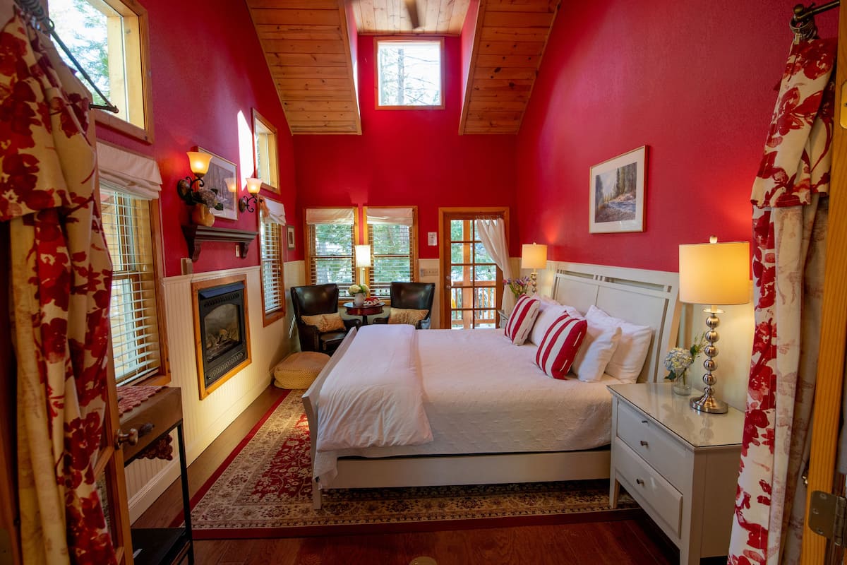 A bedroom with a large bed, a fireplace, a desk, and a window with a view of trees.