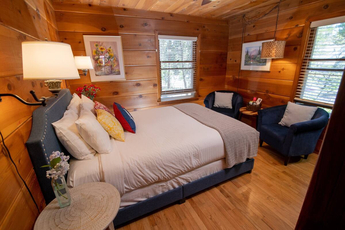 A wooden cabin bedroom with a bed, two chairs, a small table, and a window with blinds.