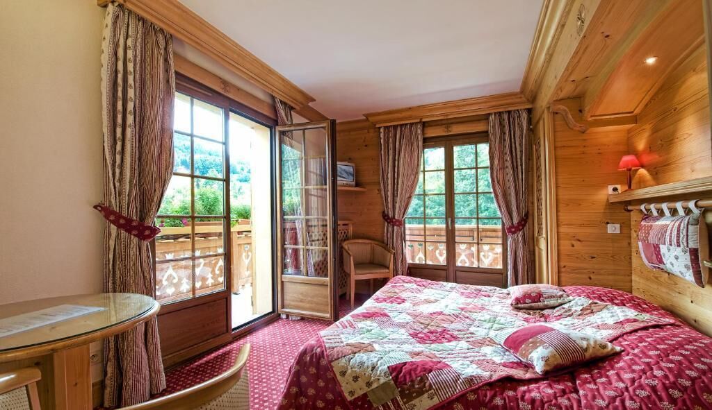 Ein Zimmer mit einem Bett, einem Sessel, einem Tisch und einem fensterbepflanzten Fenster.