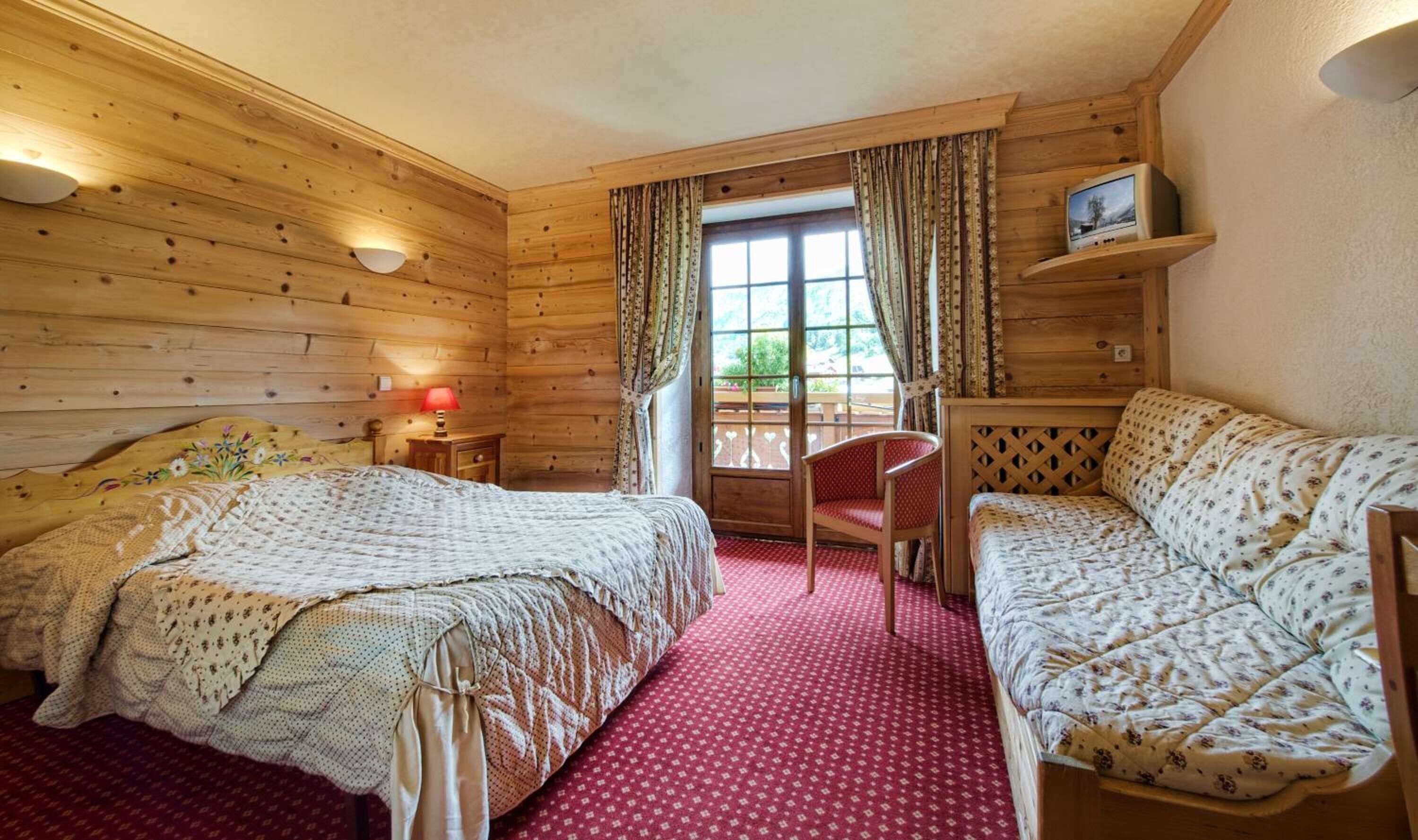 A wooden cabin bedroom with two beds, a red carpet, a TV on a shelf, and a window with curtains.