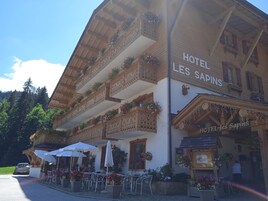 A hotel with multiple balconies adorned with flowers, outdoor seating with umbrellas, and a sign reading "HOTEL LES SAPINS".