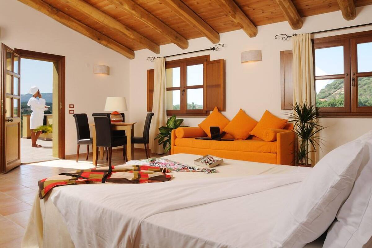 A bedroom with a bed, an orange sofa with cushions, a desk with a chair, and a view of the outdoors.