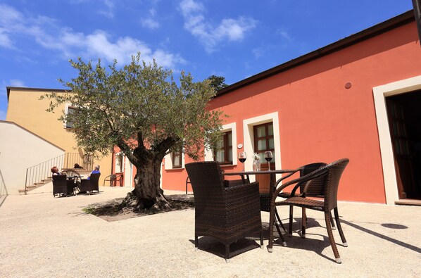 Un espacio al aire libre con sillas de mimbre, una mesa, un árbol y un edificio de fachada roja.