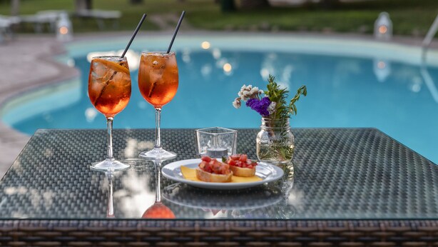 Una mesa con dos bebidas, un plato de comida y un florero con flores junto a una piscina.