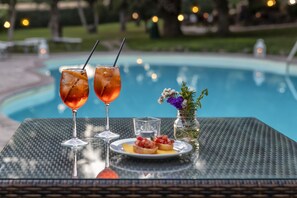 Una mesa con dos bebidas, un plato de comida y un florero con flores junto a una piscina.