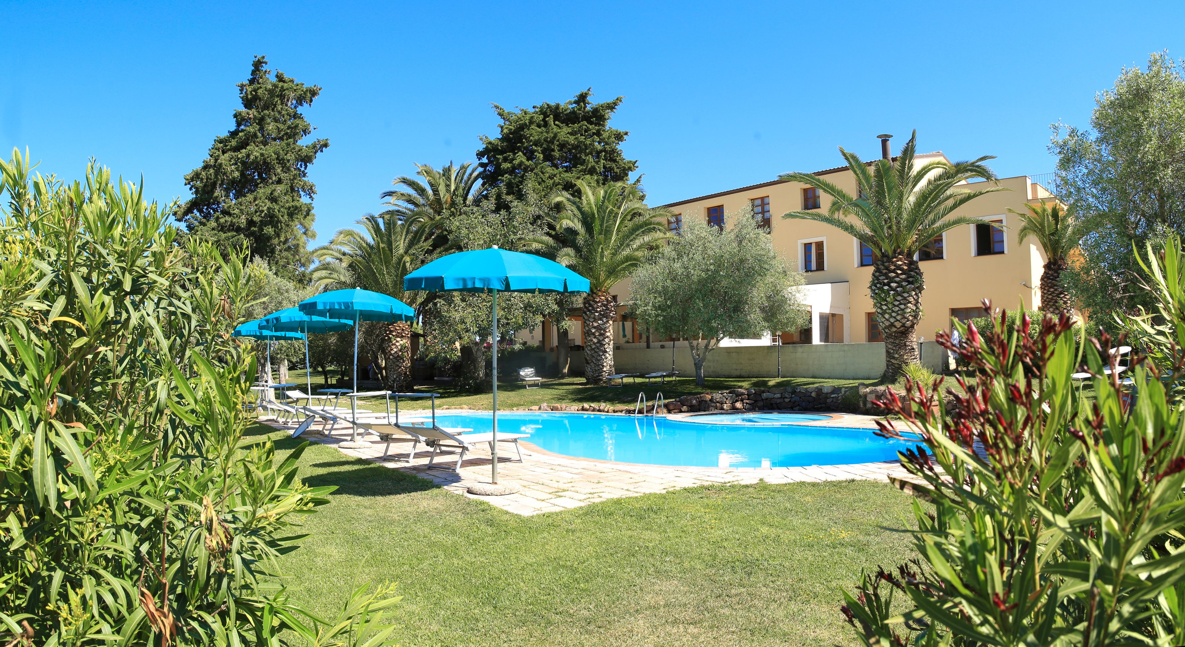 Zona de piscina con sillones de descanso y sombrillas, rodeada de follaje y un edificio al fondo.