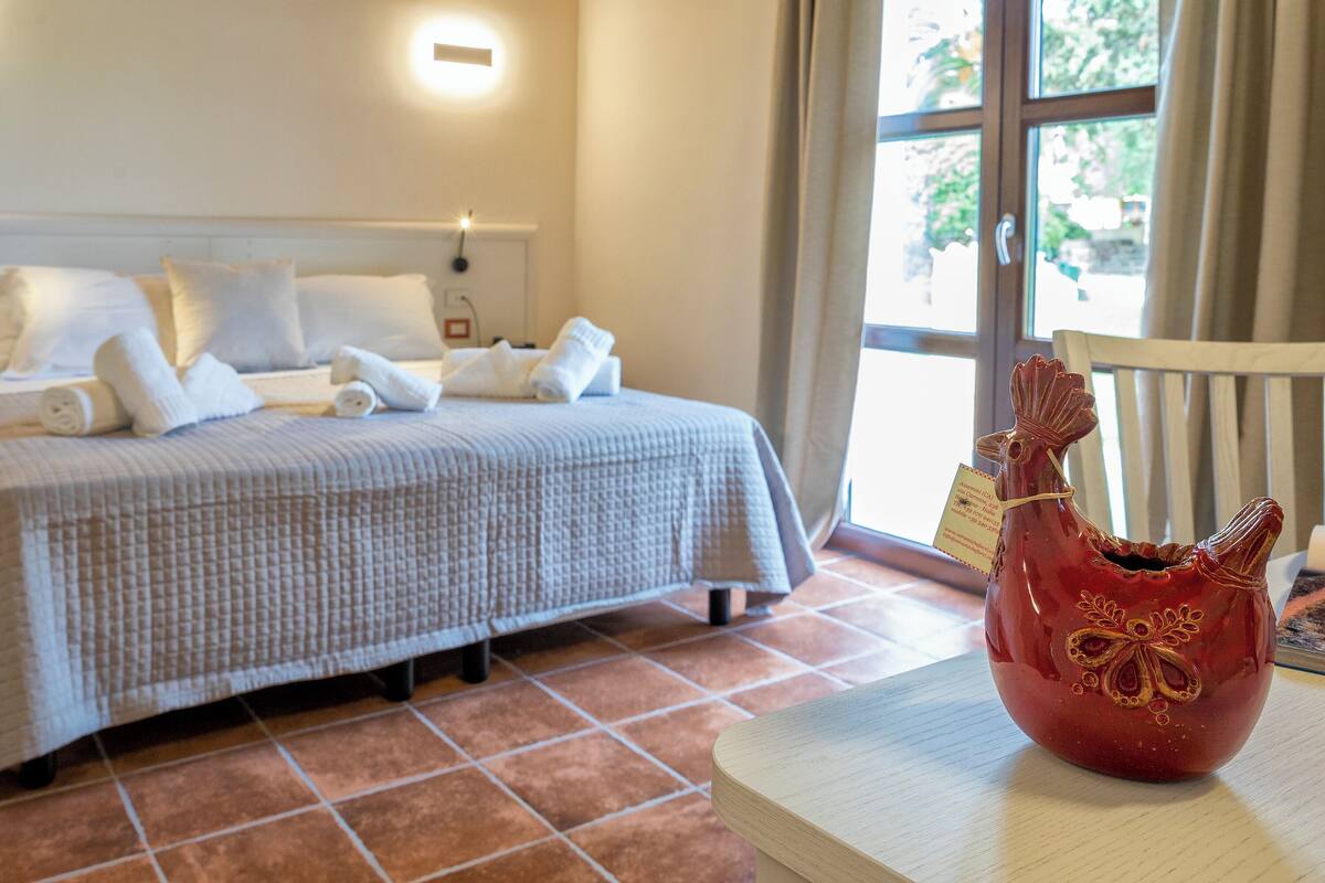 A neatly made bed with white linens and a red decorative vase on a wooden table.