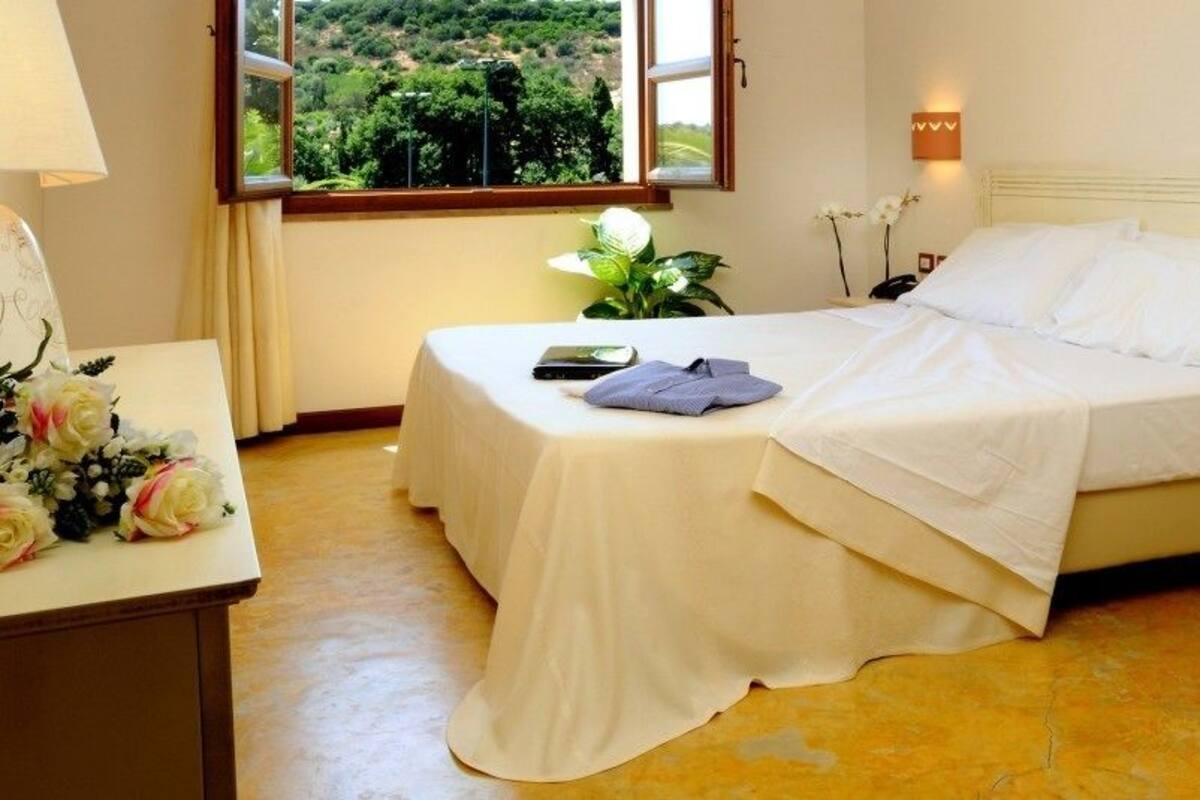A bedroom with two beds, a bedside table with a lamp, a nightstand, and a window with a view of greenery.