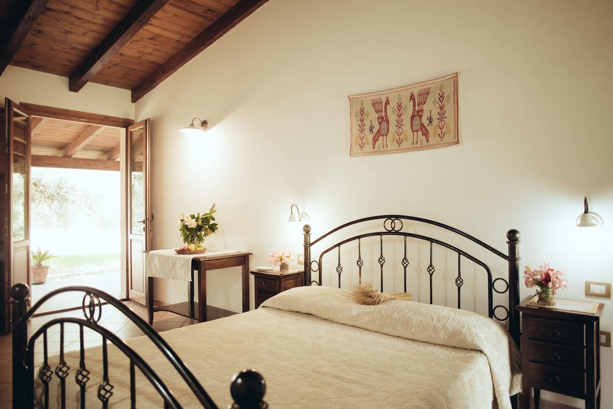 A bedroom with a bed, bedside tables, a nightstand, a wall-mounted lamp, and a window with a view of greenery.