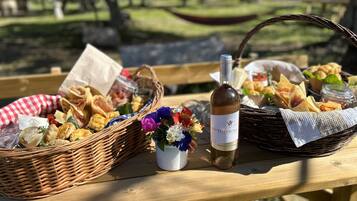 A picnic setup with baskets of food, a bottle of wine, and a small vase of flowers on a wooden table outdoors.