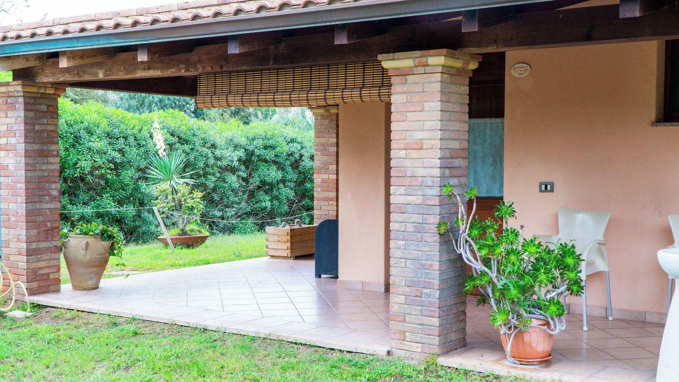 Un área de patio techada con pilares de ladrillo, plantas en macetas y suelo de baldosas.