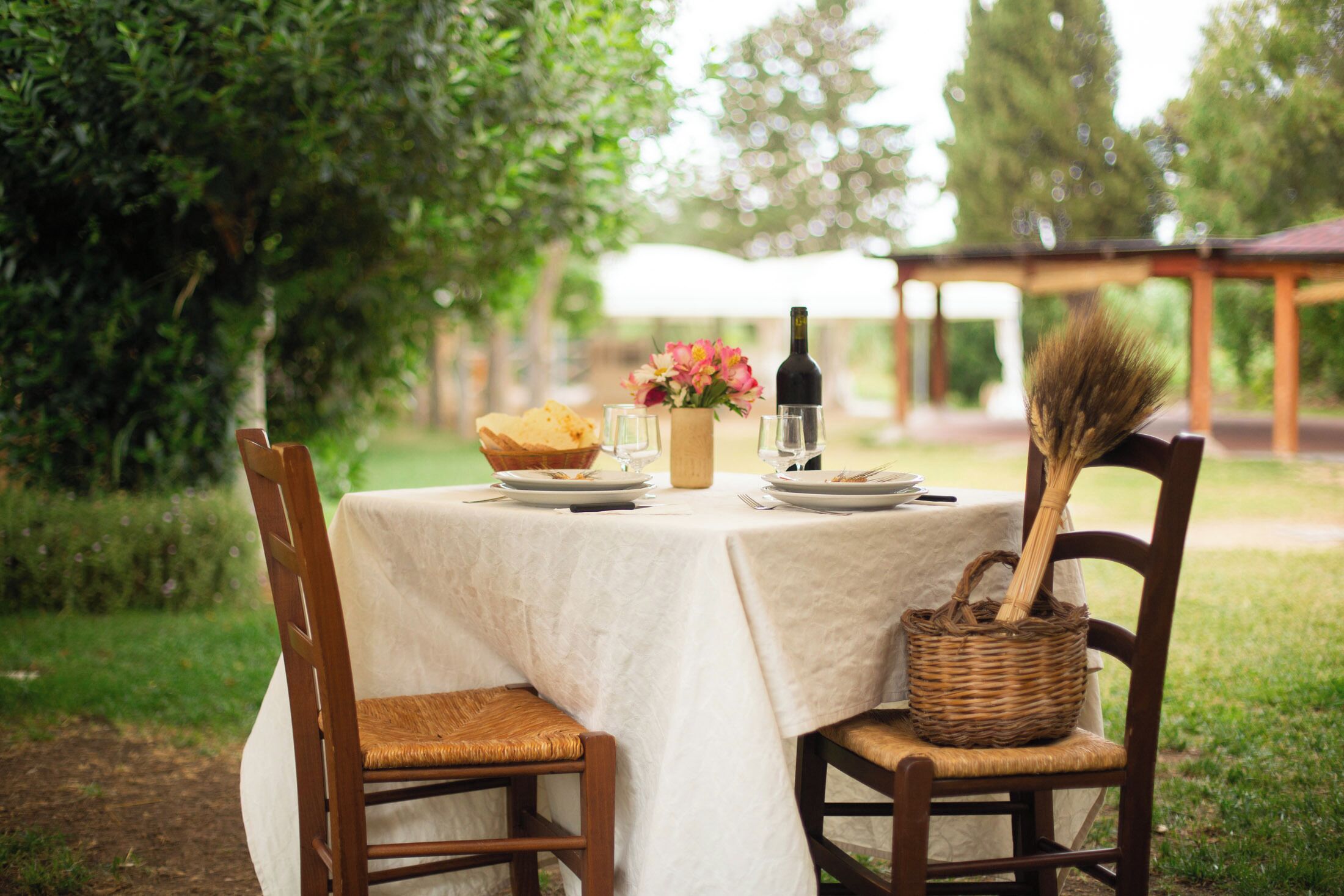 An outdoor dining table with a white tablecloth, surrounded by wooden chairs. There is a basket, a bottle, and a vase with flowers on the table.