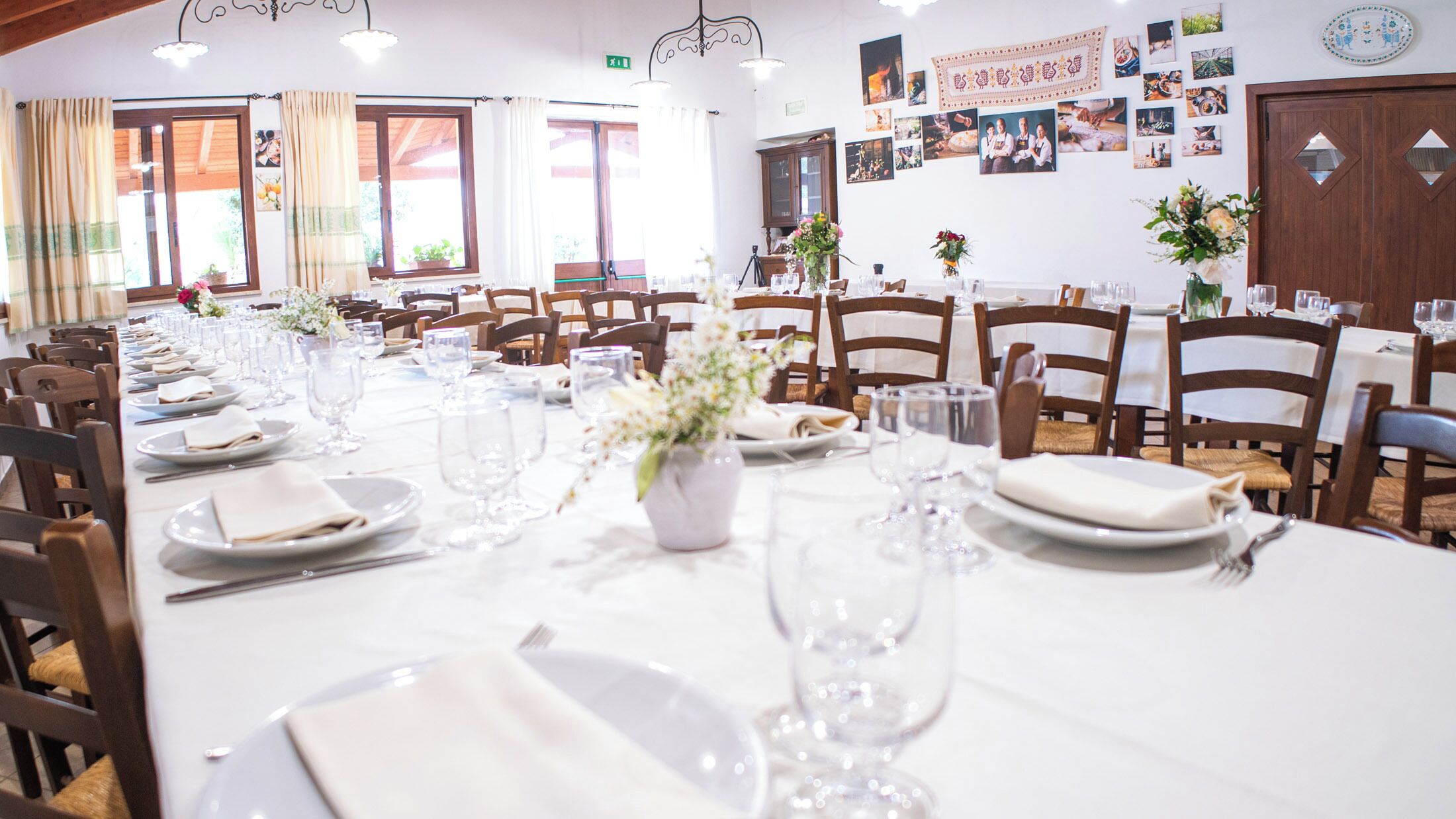 A long dining table set with white tablecloths, plates, and wine glasses, surrounded by wooden chairs.