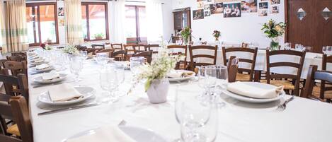A long dining table set with white tablecloths, plates, and wine glasses, surrounded by wooden chairs.