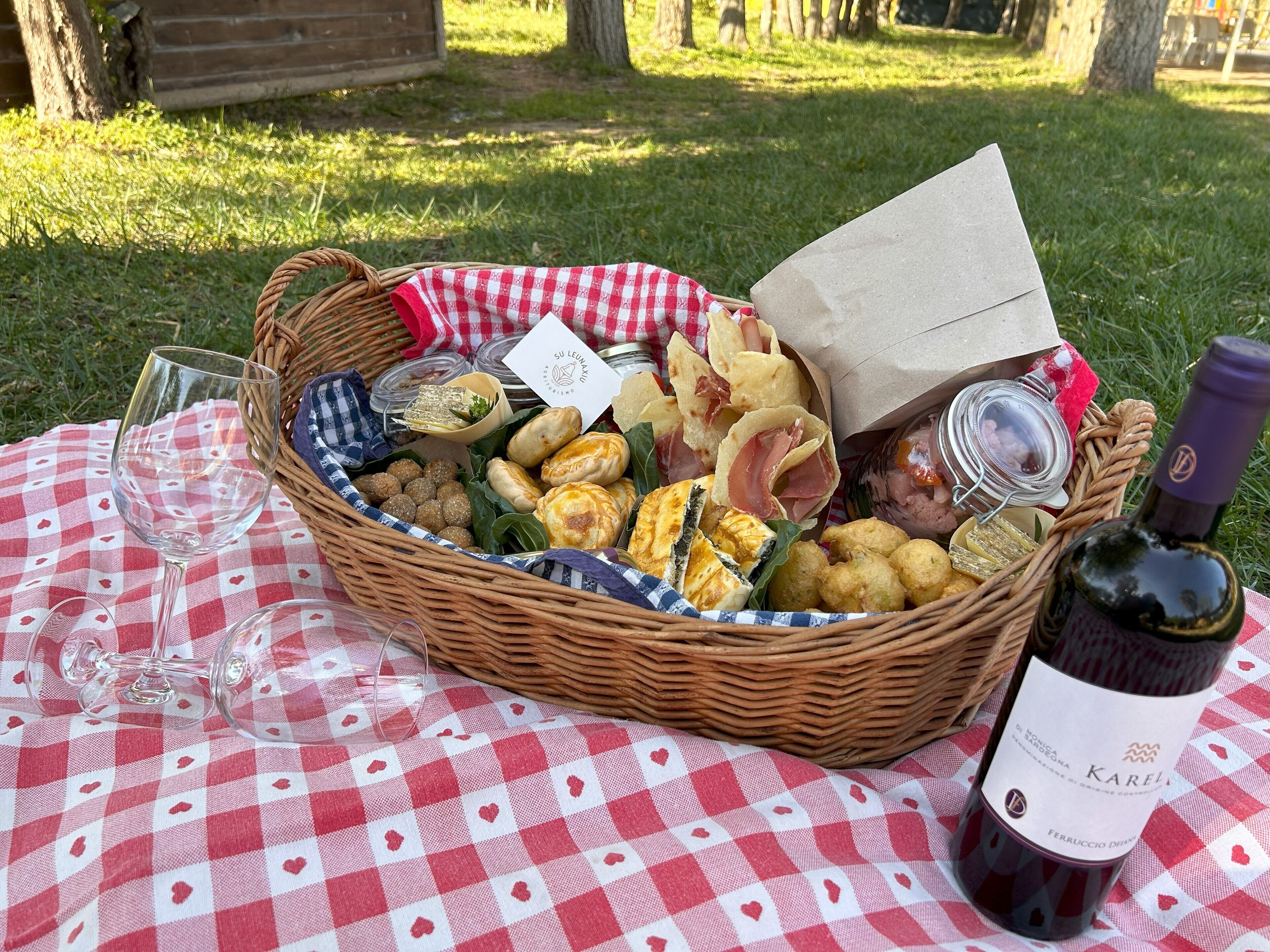 Uma cesta de vime montada para piquenique, com comida, uma garrafa de vinho e um copo sobre uma toalha xadrez.