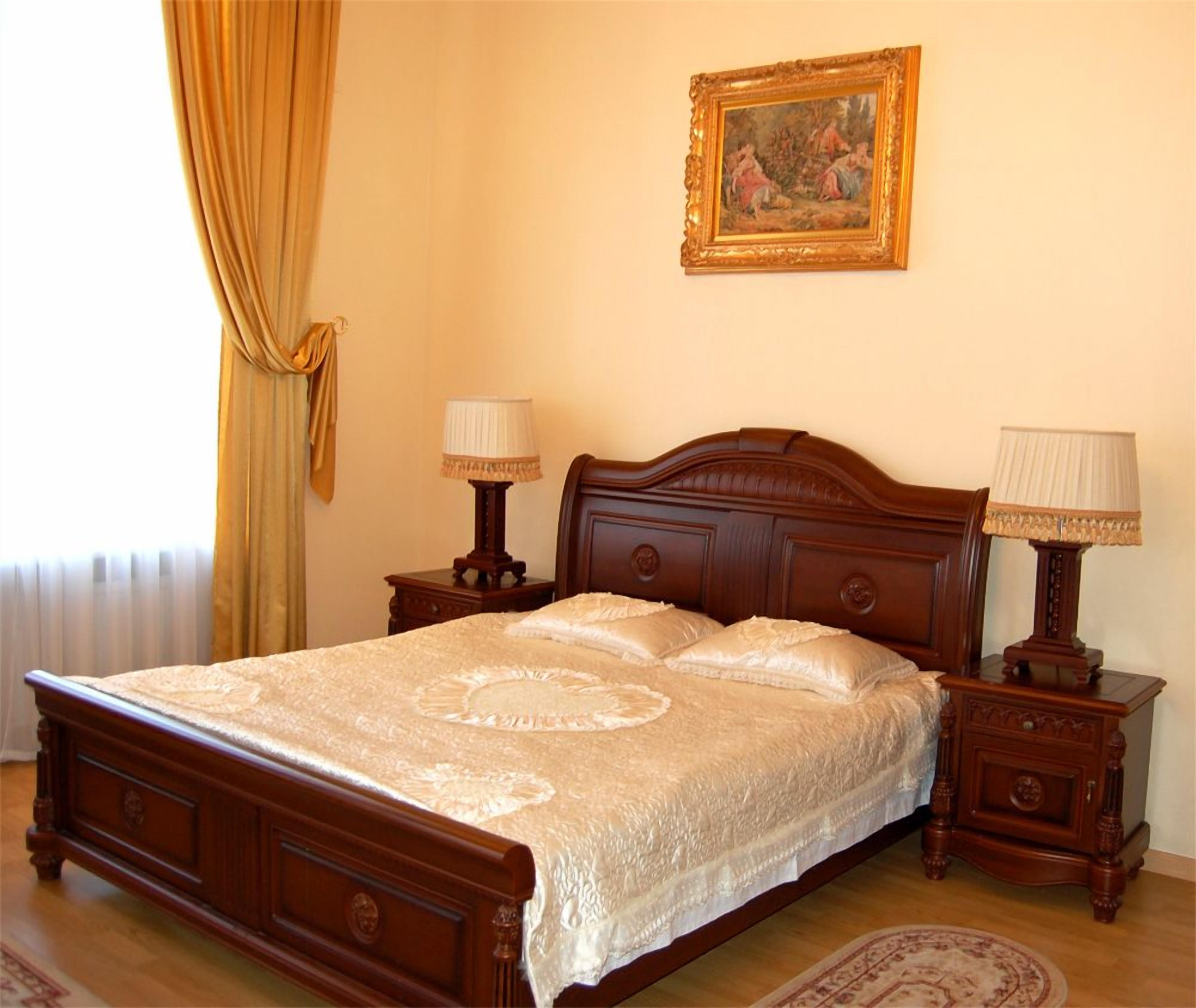 A bedroom with a wooden bed, two bedside tables with lamps, a framed painting on the wall, and a window with curtains.